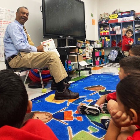 This is an image of a man reading a book to students in a classroom.