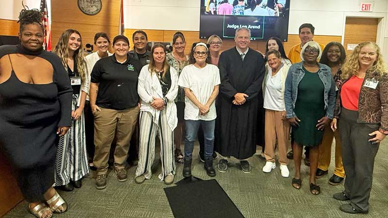 This is a group of people in the front of a courtroom standing with a judge dressed in a black robe.