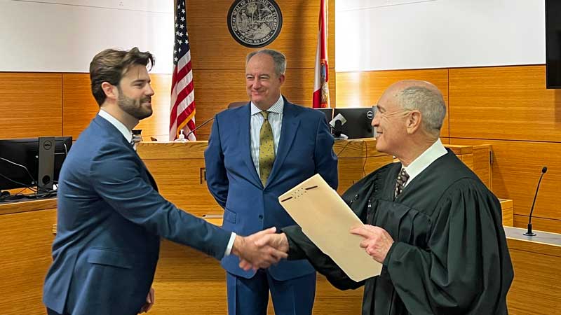 This is an image of a man dressed in a black robe shaking the hand of another man in the front of a courtroom.