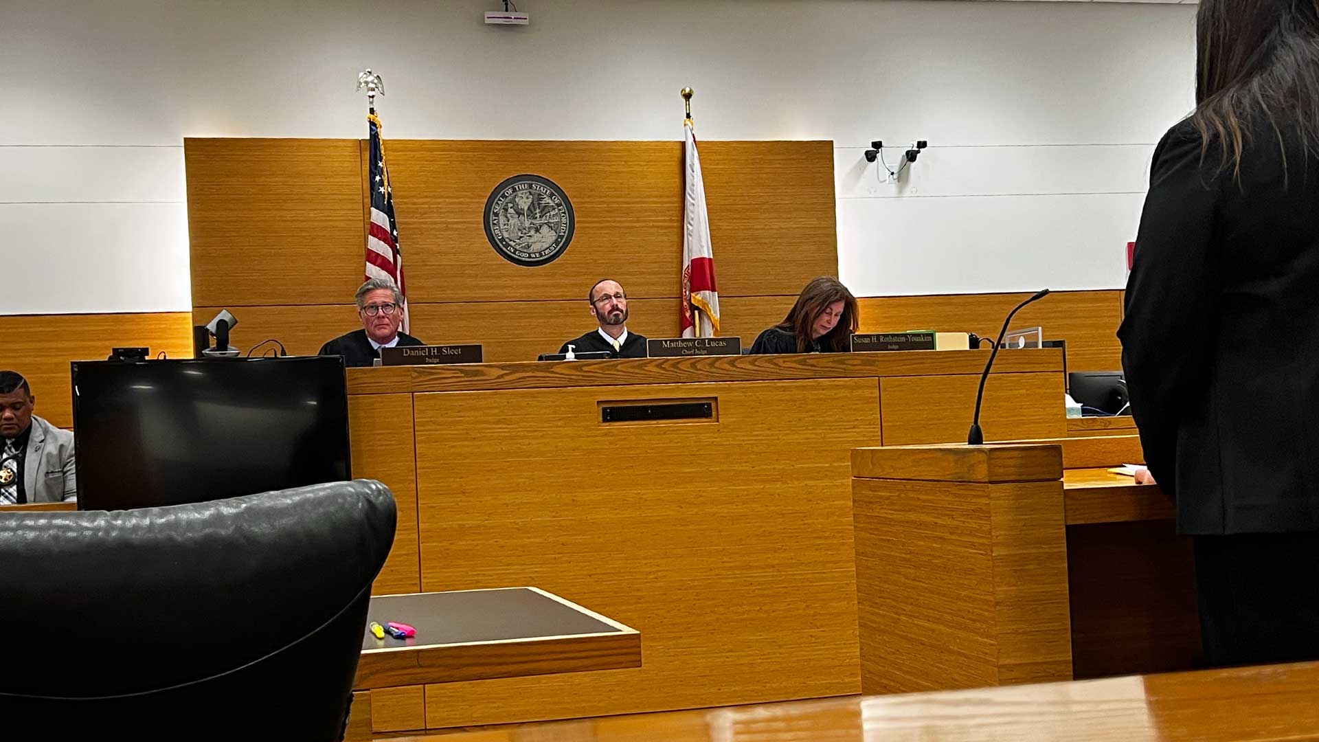 This is a photograph of three judges, two men and one woman, sitting in a courtroom listening to a lawyer speak from a lectern in front of them.