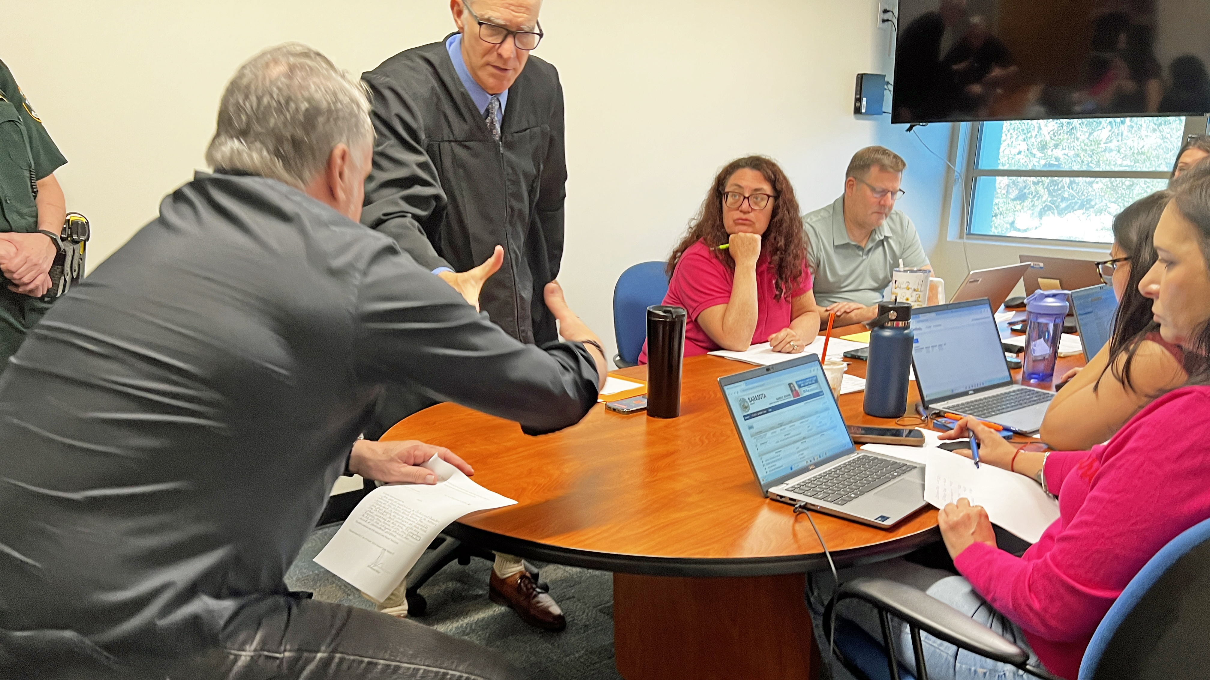 This is a photograph of a male judge dressed in a black robe reaching to shake the hand of a man seated at a table. There are five other people -- four men and one woman -- seated at the table, some watching the judge, others looking at computer screens.