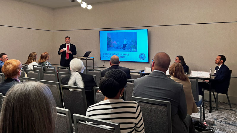A mam at the front of the room speaks to a group of people while showing a slideshow presentation.